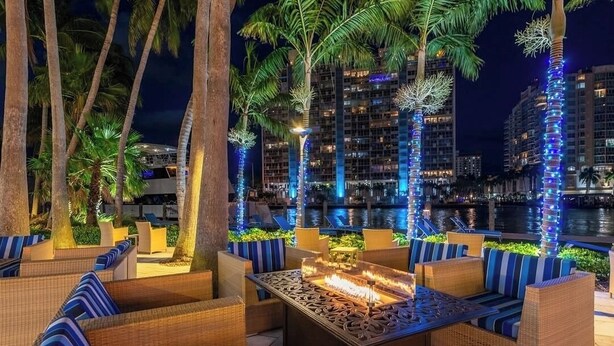 Zona de estar al aire libre con mobiliario de mimbre y un hogar central, rodeada de palmeras e iluminada con luces azules.