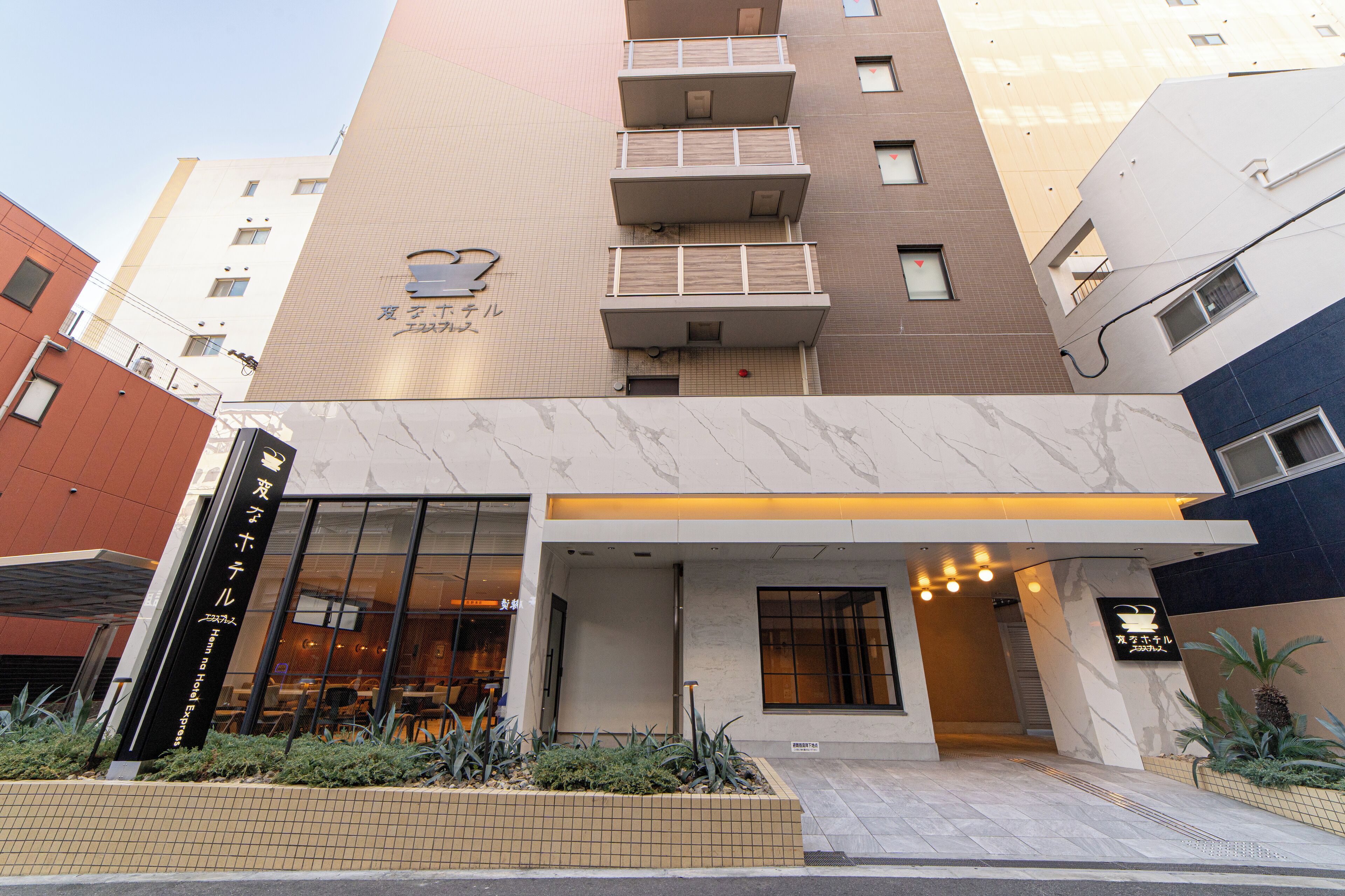 A hotel entrance with a tiled facade, a visible signboard, and a small garden area with plants.