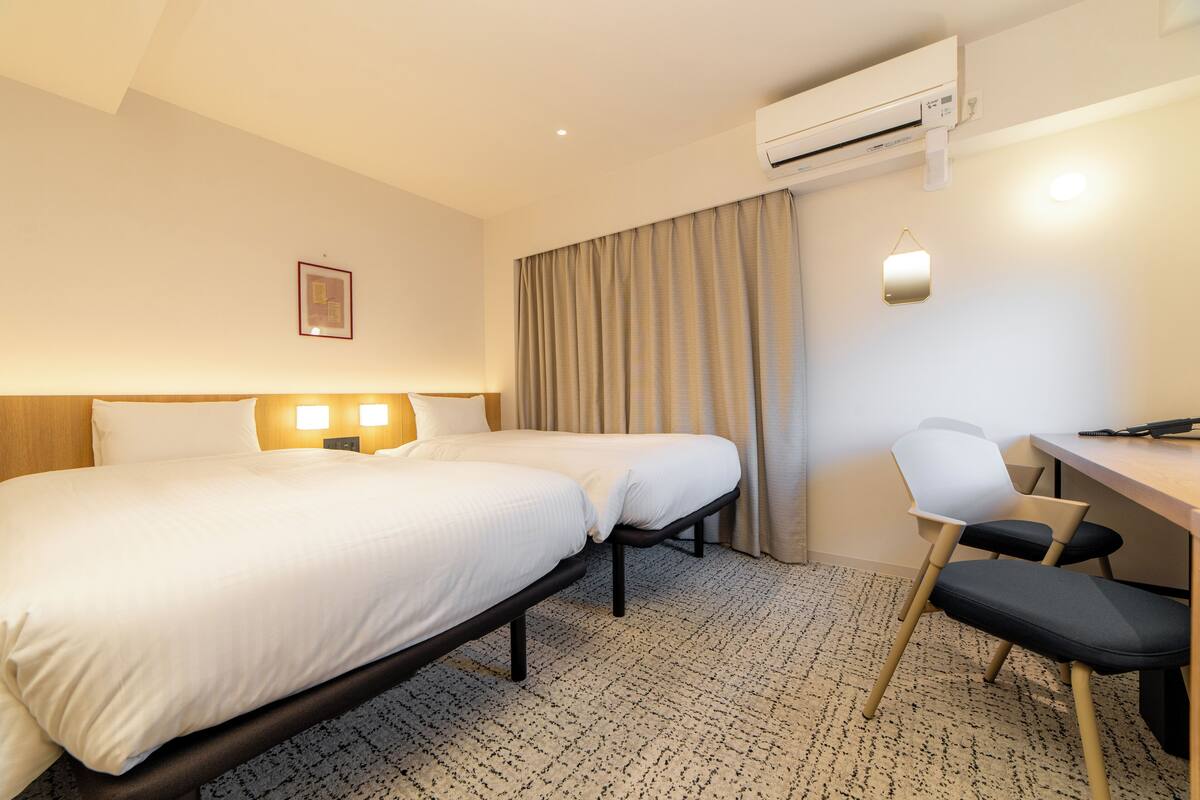Une chambre d’hôtel avec deux lits, un bureau, une chaise, un appareil de climatisation et un luminaire fixé au mur.