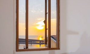 Une fenêtre donnant sur un paysage où l’on aperçoit un coucher de soleil sur une étendue d’eau, un pont et un bâtiment avec un toit recouvert de tuiles.