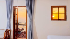 A room with a bed, a wooden chair, a table with chairs, a window with a view of the sunset, and curtains. - Sentina Phu Quoc Hotel (Phu Quoc)