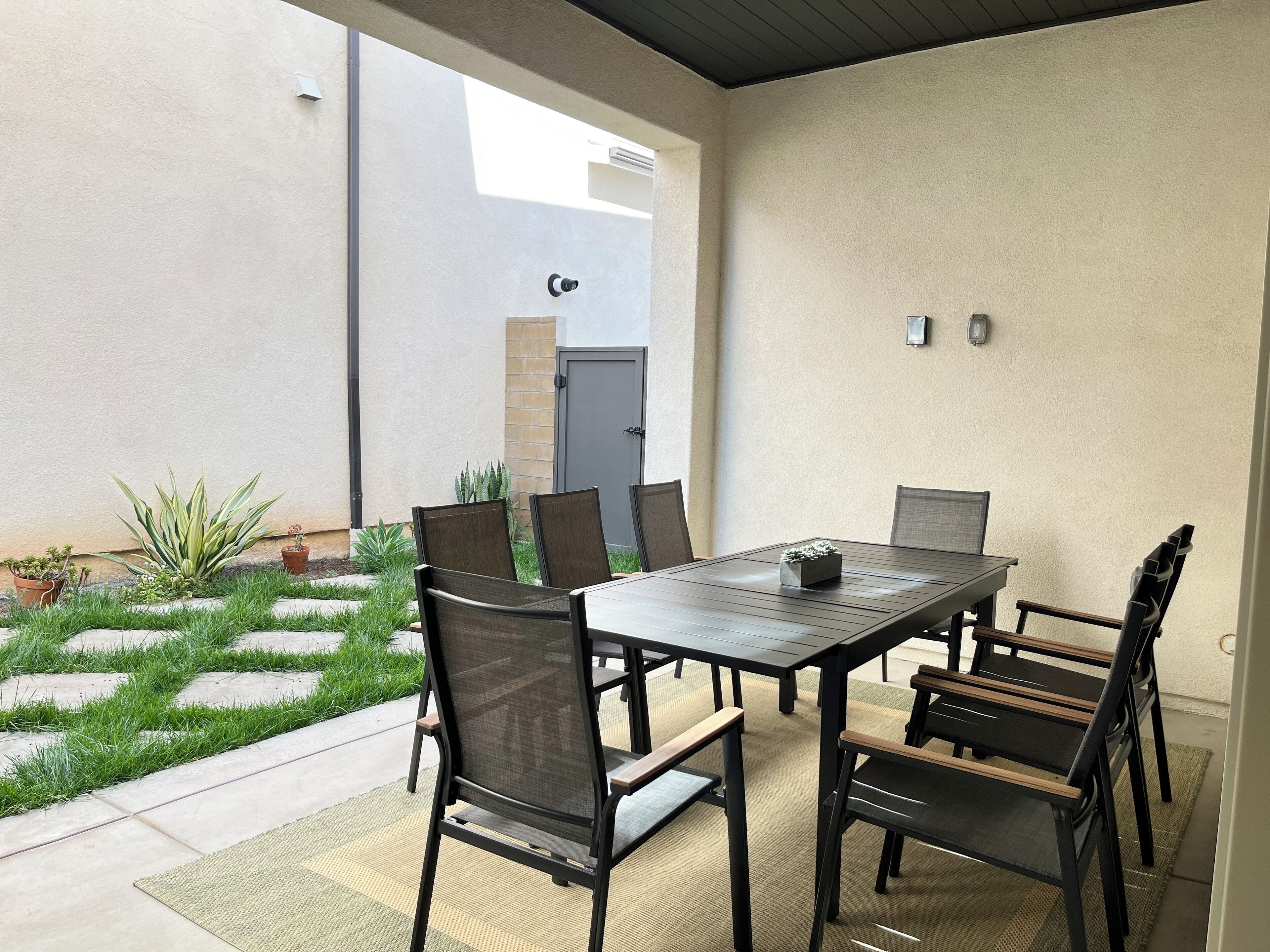 An outdoor patio with a rectangular table and six chairs, surrounded by a small garden with grass and potted plants.
