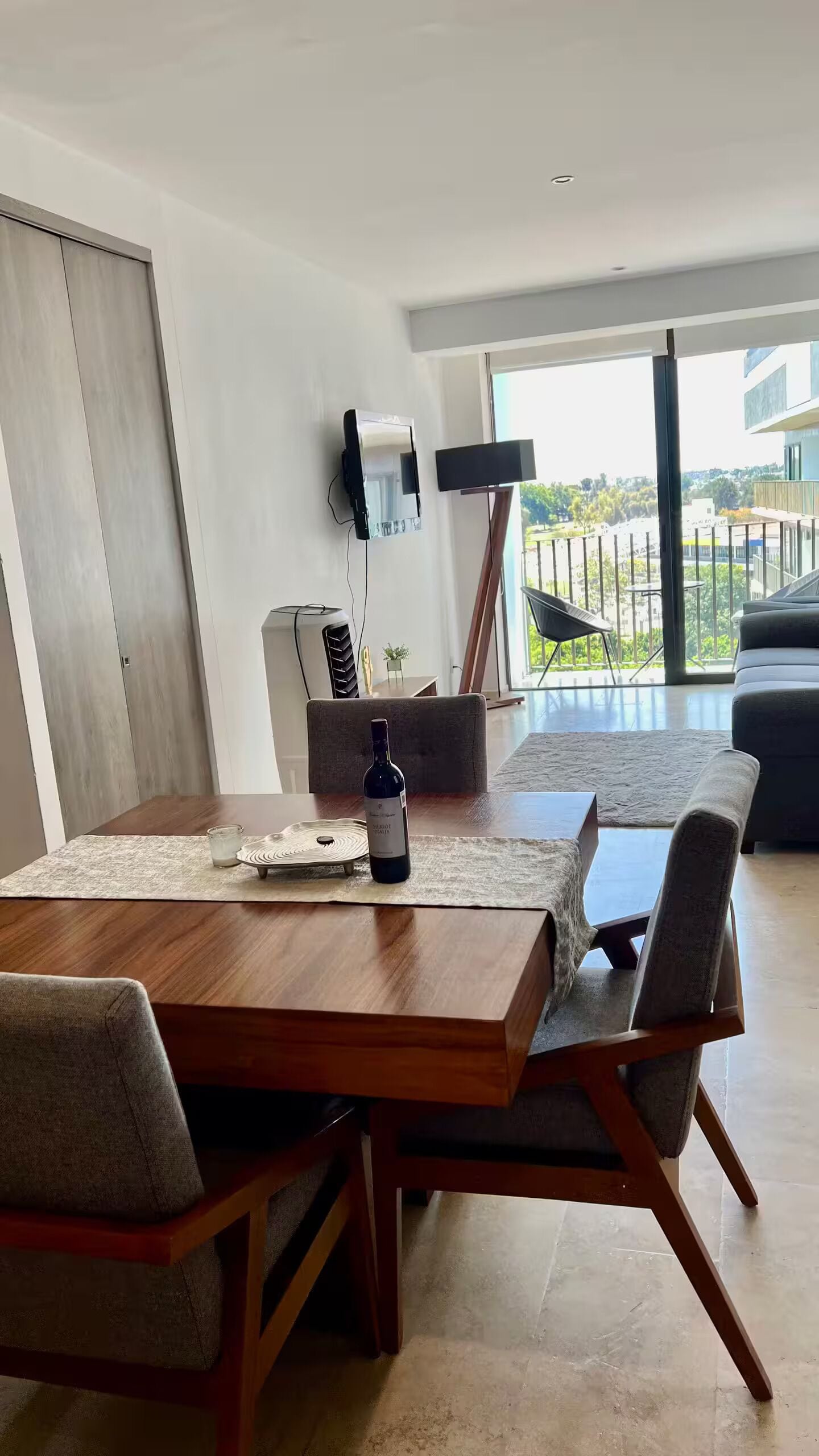 Ein moderner Essbereich mit einem Holztisch, grau gepolsterten Stühlen und einer Weinflasche. Ein Flachbildfernseher ist an der Wand montiert, und ein großes Fenster bietet Blick ins Freie.