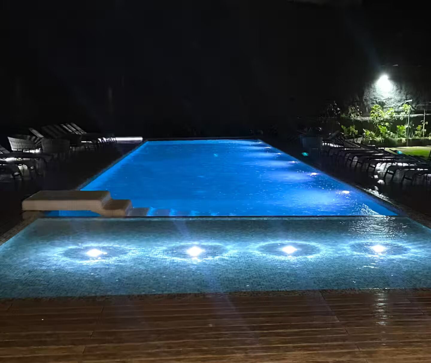 A swimming pool with a wooden deck in the foreground and lounge chairs on the left.
