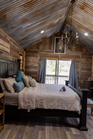A rustic wooden bedroom with a large bed, a chandelier, and a view through a window. - Upper Room - Come explore the wild wonders of Oak Meadow Ranch (Valley View)