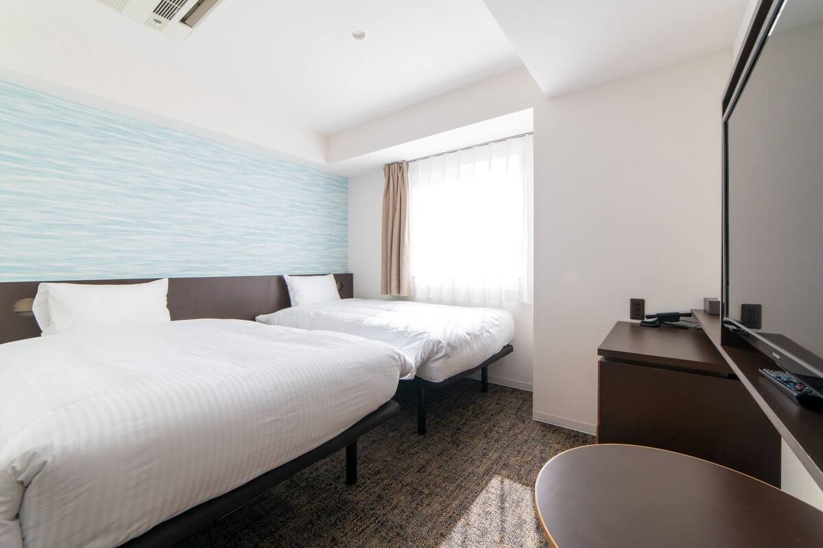 Une chambre d’hôtel avec deux lits, un bureau, une chaise, un grand miroir et une fenêtre avec des rideaux.
