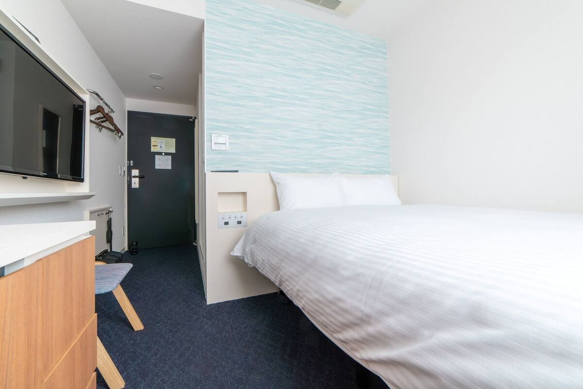 Une chambre d’hôtel avec un lit, un téléviseur fixé au mur, une commode en bois et une porte donnant sur une autre pièce.