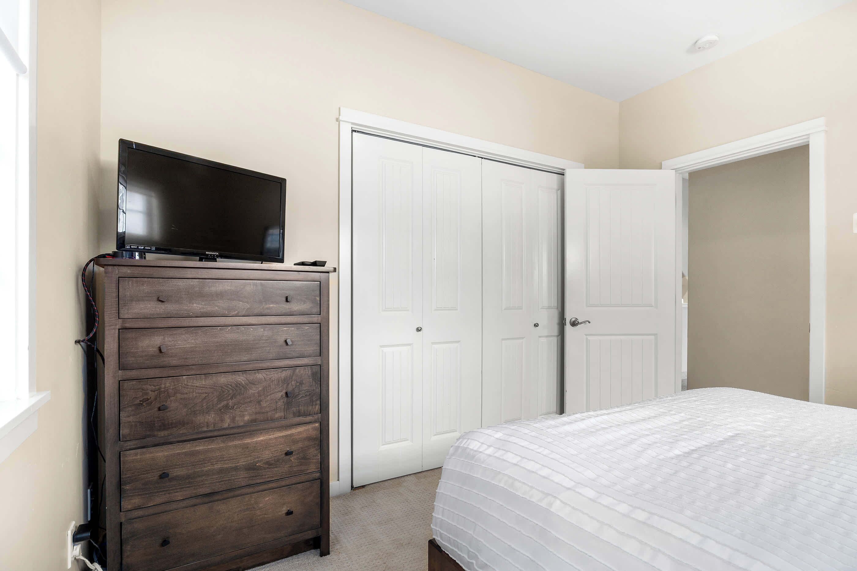 Een slaapkamer met een houten dressoir, een televisie erboven, een wit bed met een witte spreidek en twee witte deuren.