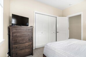 Un dormitorio con un aparador de madera, un televisor encima, una cama blanca con colcha blanca y dos puertas blancas.