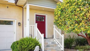A house with a red door, white steps, and a small garden with a tree and shrubs.
