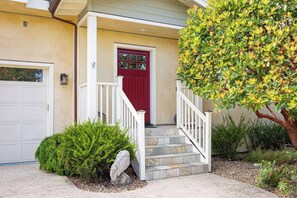 A house with a red door, white steps, and a small garden with a tree and shrubs.