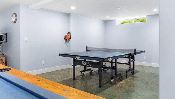 A ping pong table in a room with light blue walls, a clock on the wall, and a small window with greenery visible.