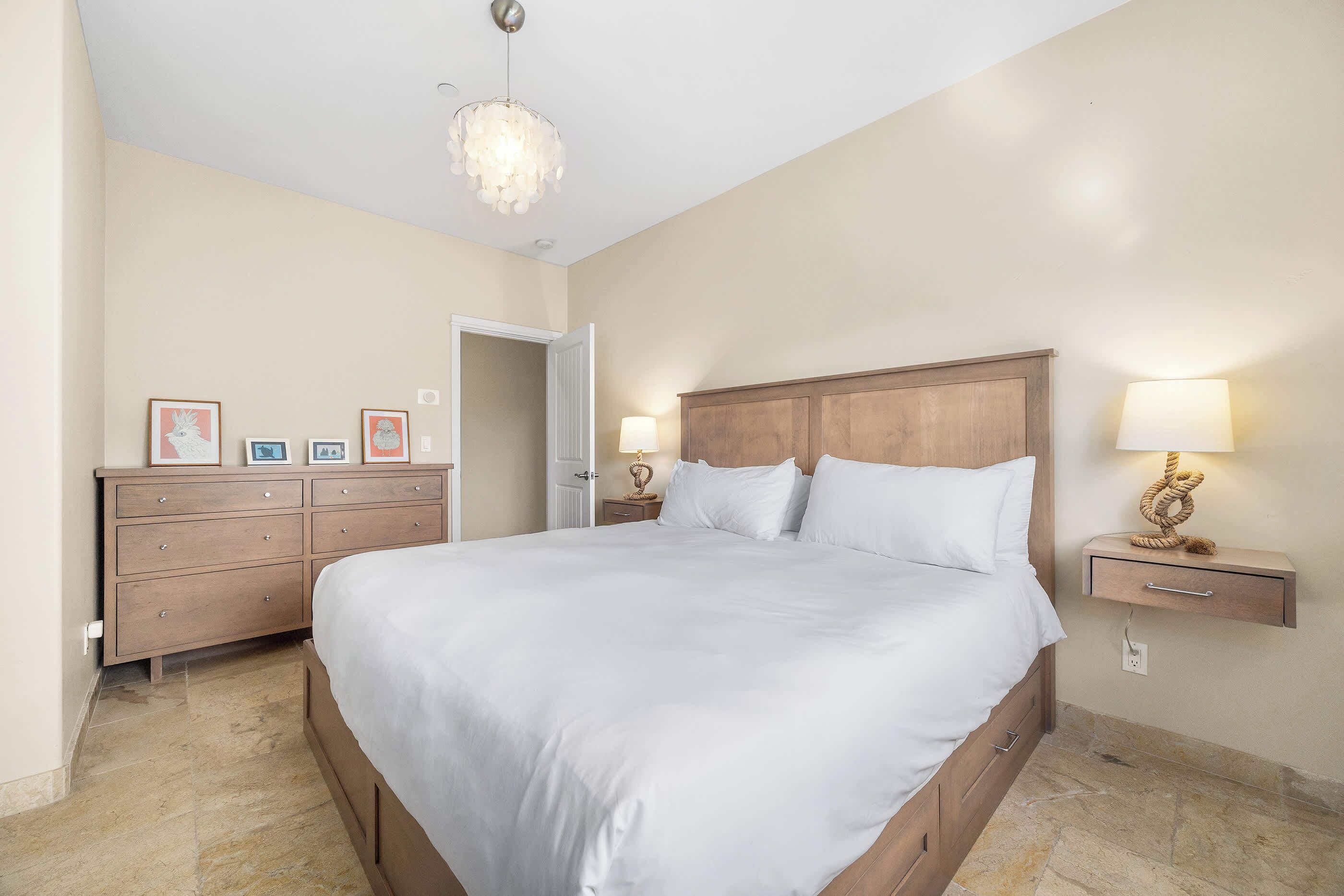 A bedroom with a large bed, wooden headboard, bedside tables with lamps, a dresser, and framed pictures on the wall.