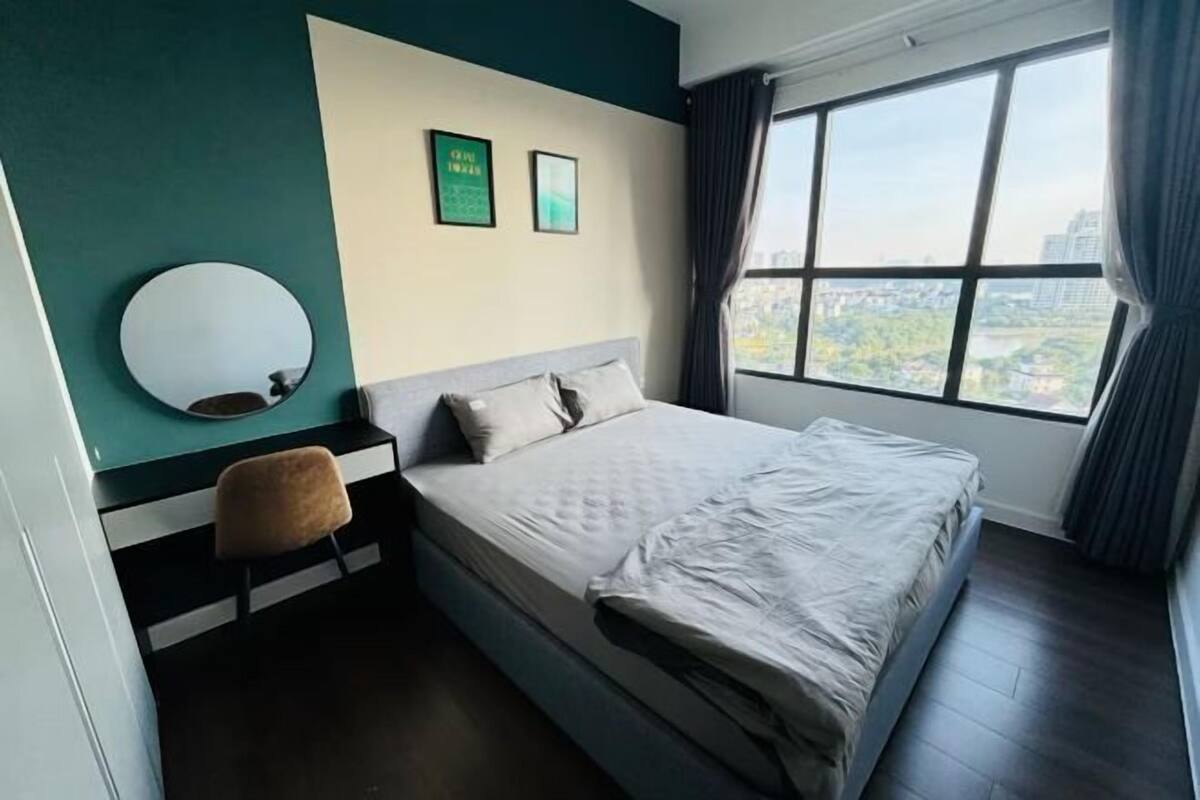 A hotel room with a bed, a chair, a round mirror, and a window with a city view.