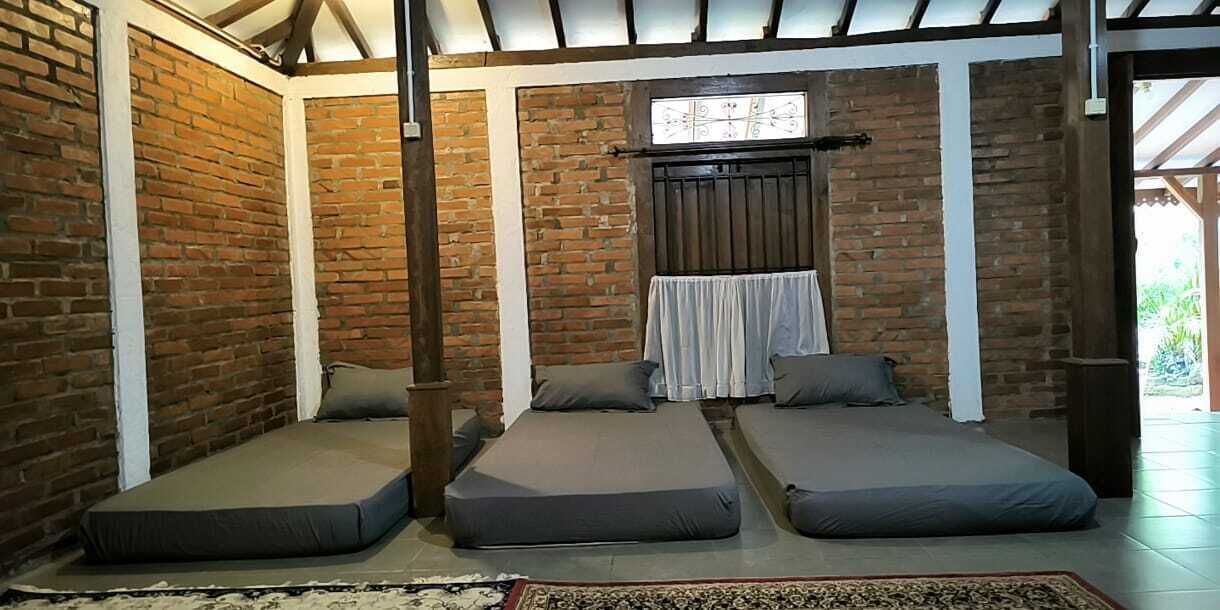 A room with three single beds aligned against a brick wall, a window with a curtain, and a wooden beam ceiling.