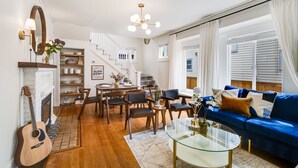 Ein Wohnzimmer mit einem blauen Sofa, einem Glastisch, einem Kamin, einer Gitarre und einer Treppe, die zu einem Schlafboden führt.