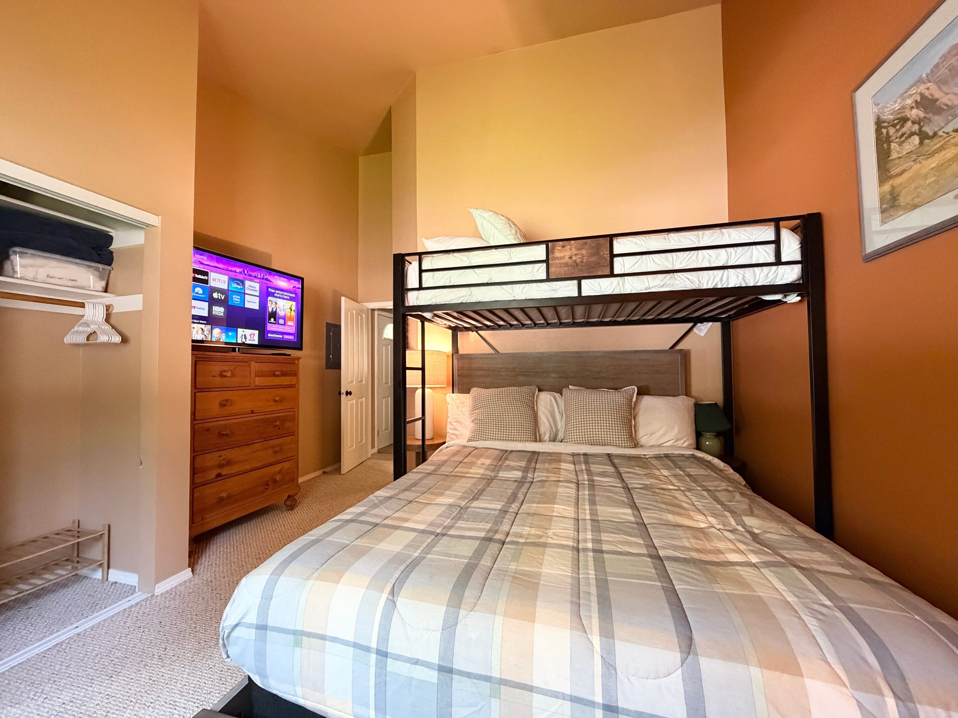 Un dormitorio con una cama litera, un televisor sobre un mueble bajo, un aparador de madera y una puerta que da a otra habitación.
