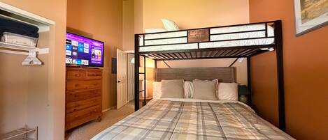 Un dormitorio con una cama litera, un televisor sobre un mueble bajo, un aparador de madera y una puerta que da a otra habitación.
