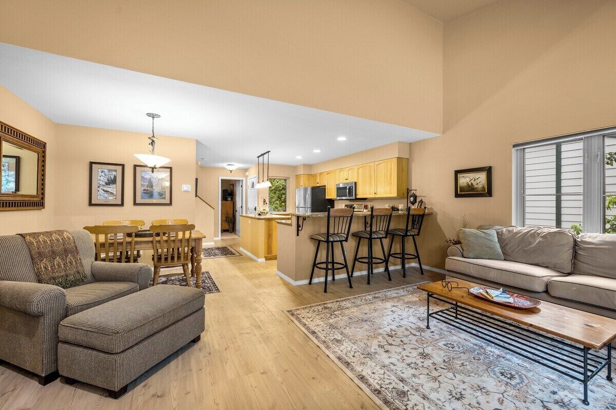 A modern living room with a sofa, ottoman, and coffee table, a dining area with chairs and a table, and a kitchen with bar stools.