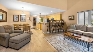 A modern living room with a sofa, ottoman, and coffee table, a dining area with chairs and a table, and a kitchen with bar stools.