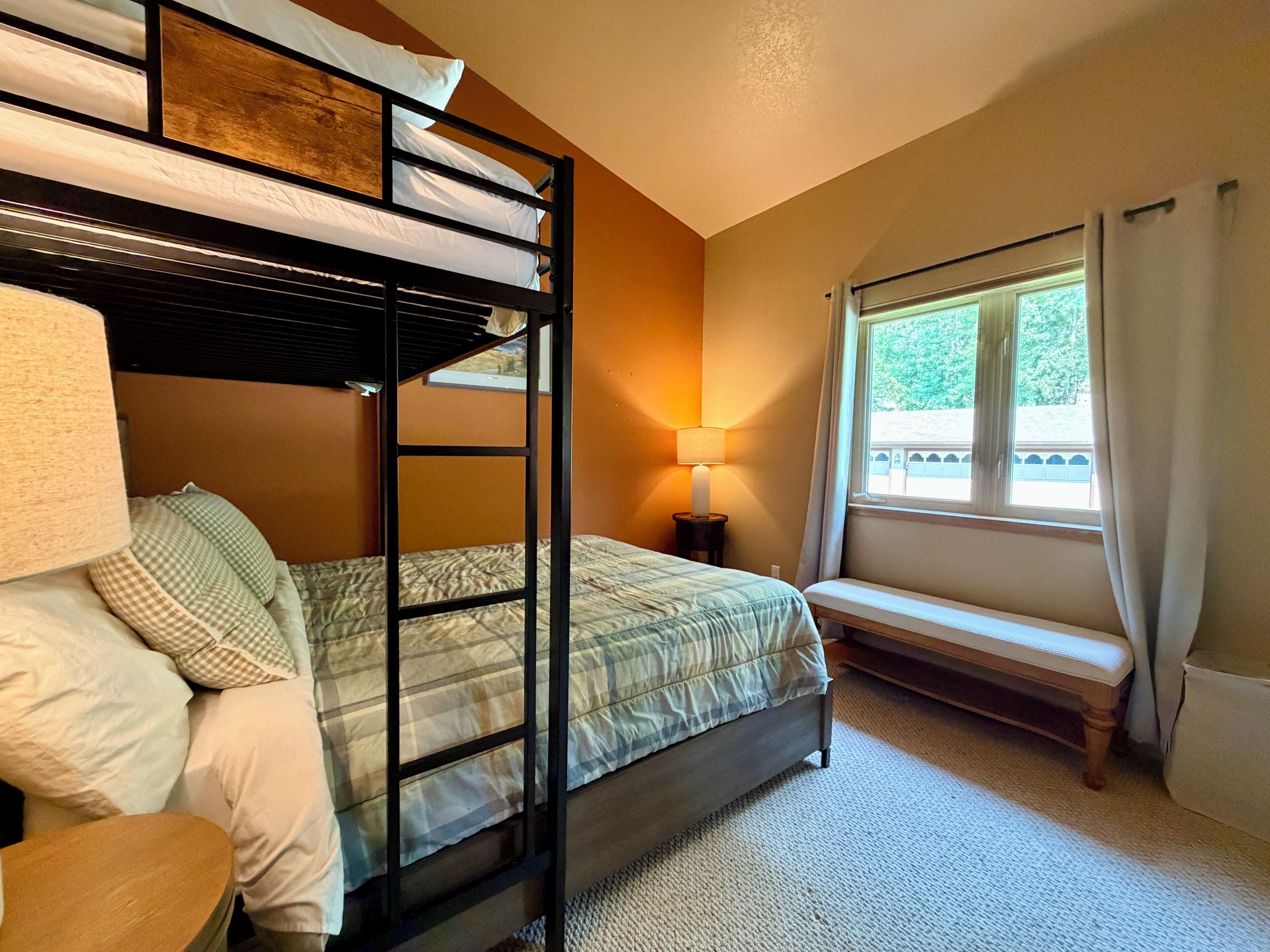 A bunk bed room with a window, a bench, and a lamp.