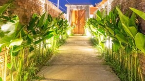 A pathway flanked by brick walls and lush green plants leading to a wooden door.
