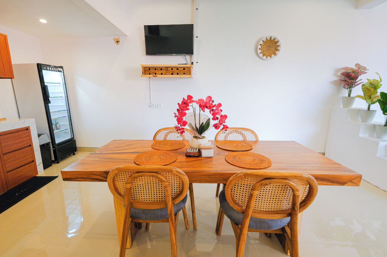 Un coin repas avec une table en bois, quatre chaises en osier, un vase avec des fleurs rouges, un téléviseur fixé au mur et une étagère avec des plantes.