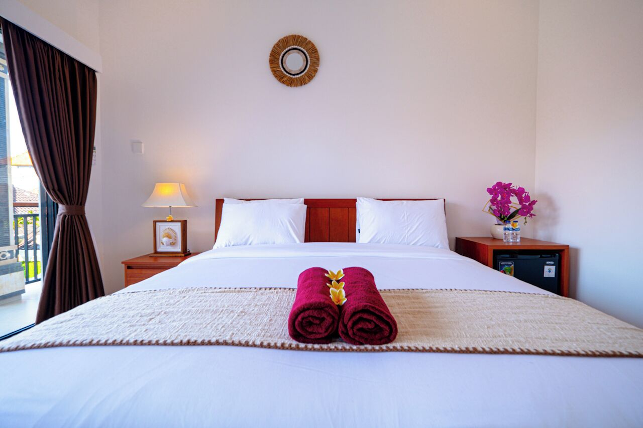 A neatly made bed with white linens, a wooden headboard, and a pair of red towels with a yellow flower.