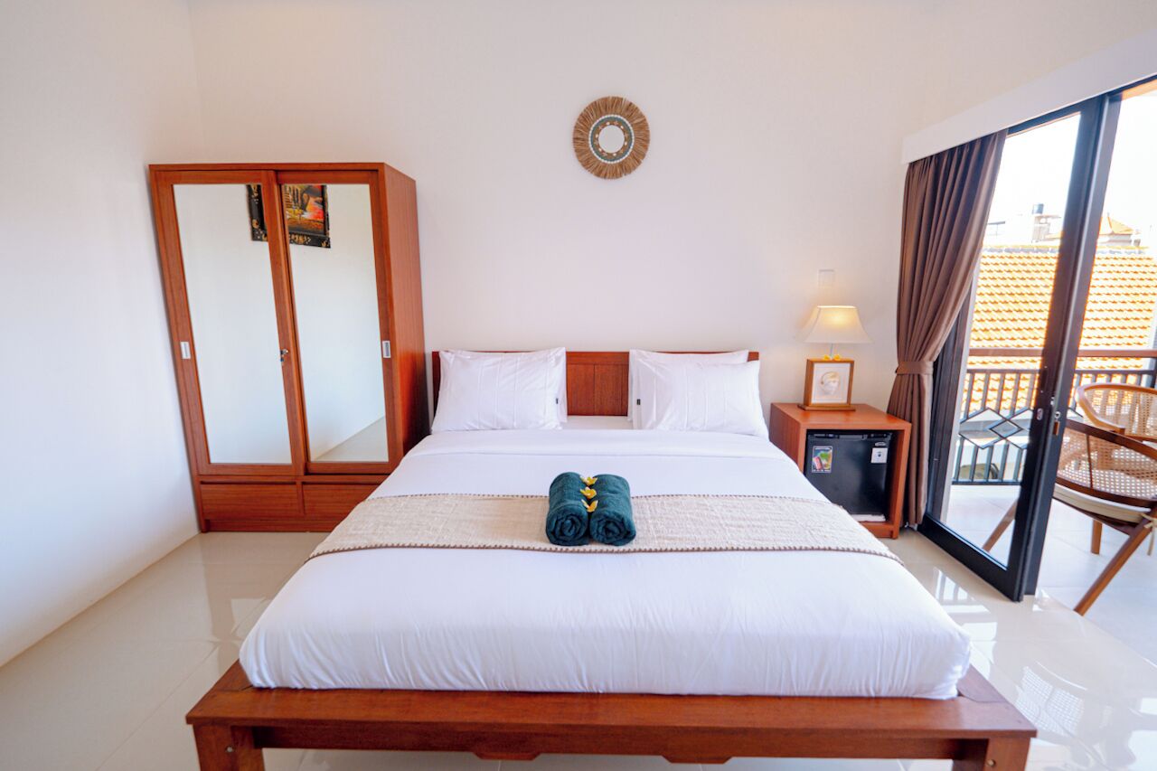 Una habitación de hotel con una cama grande, un armario de madera, una mesita de noche pequeña con una lámpara y un balcón con una silla.