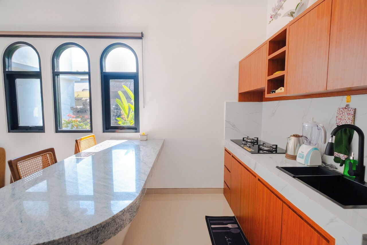 Una cocina con encimera de mármol, armarios de madera y un lavamanos negro. Hay tres ventanas con arcos y vistas al verdor exterior.