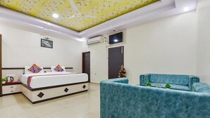 A bedroom with a bed, a blue sofa, a ceiling fan, and a TV mounted on the wall. - Hotel Gulab Vatika (Jaipur)