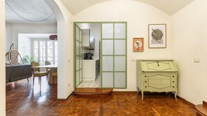 Une pièce dotée d’un parquet en bois, d’un coin repas avec des chaises et une table, d’une porte coulissante à cadre vert donnant sur une cuisine, et d’une petite commode verte avec des tiroirs.