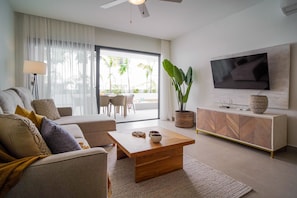 A modern living room with a sofa, coffee table, TV, and a large window with a view of palm trees. - Luxury 3 Bedroom Apartment at the Beach (Punta Cana)