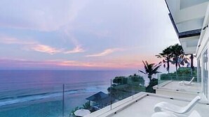 Un balcon moderne d’hôtel, doté de chaises longues blanches, d’une rambarde en verre et offrant une vue sur l’océan au coucher du soleil.