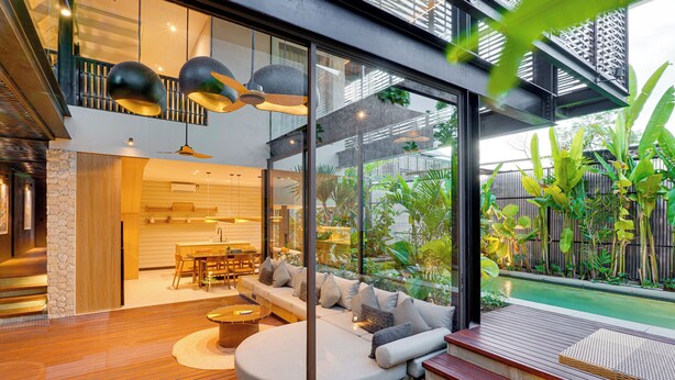 Ein modernes Interieur mit Holzboden, einer gebogenen Sitzecke mit Kissen und einer Glaswand mit Blick auf Garten und Pool.