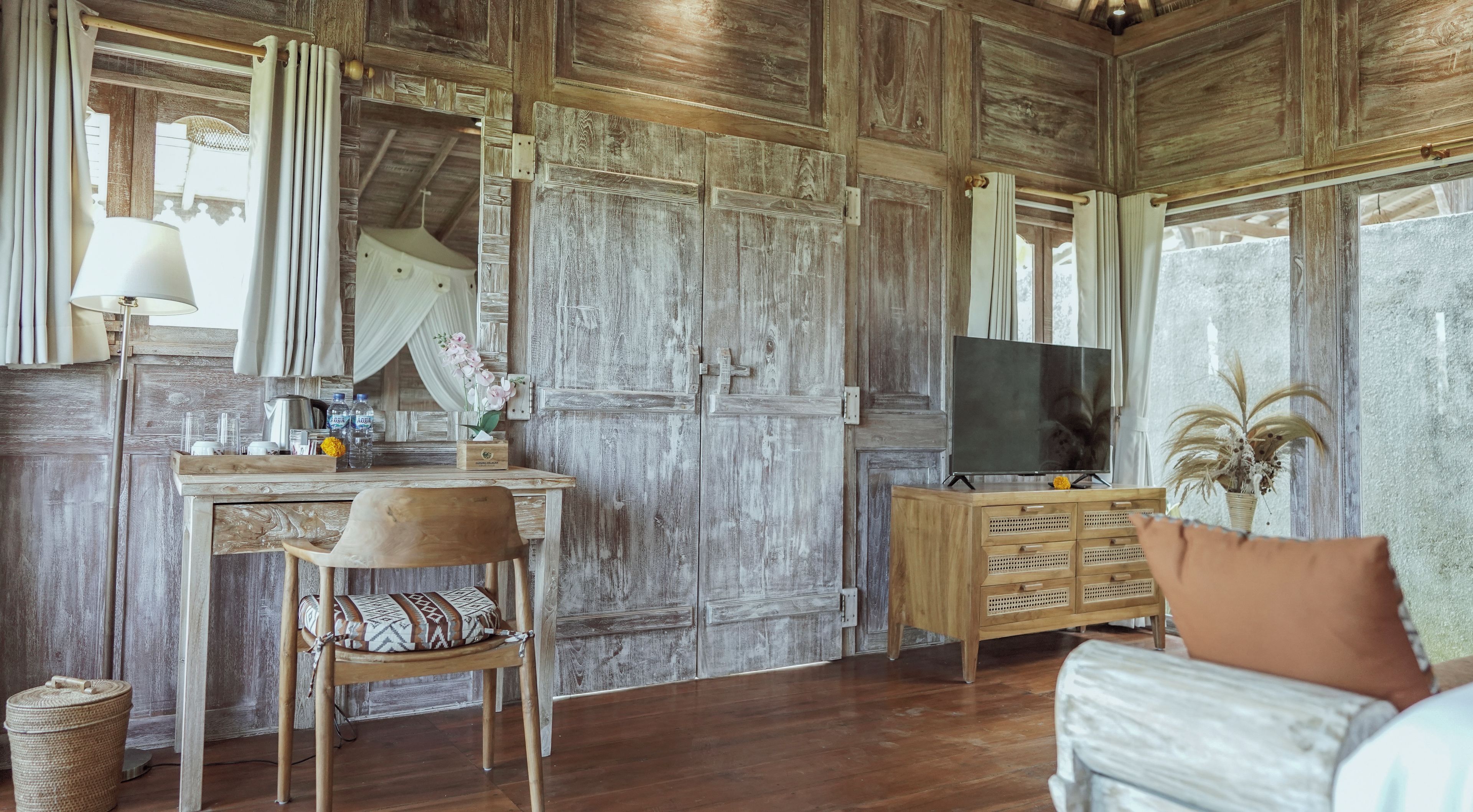 Una habitación rústica con un escritorio de madera, una silla, un televisor sobre un mueble de madera y un ventanal con cortinas.