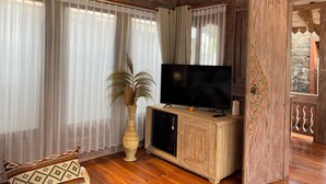 A wooden-paneled room with a television on a cabinet, a large potted plant, and sheer curtains. - Gunung Gelagah Ubud Villas by EPS (Tegallalang)