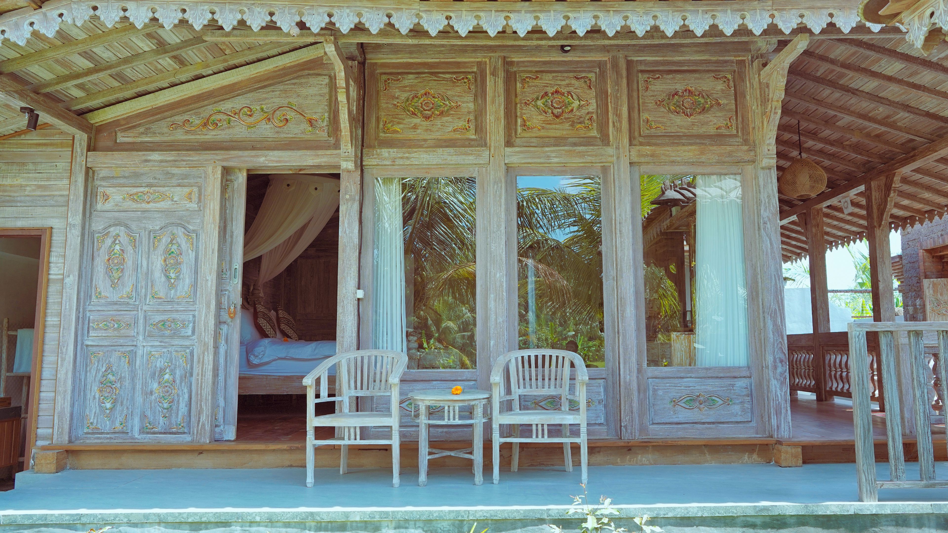Una estructura de madera tradicional con elaboradas tallas, una cama con dosel y dos sillas junto a una ventana con vistas a follaje.
