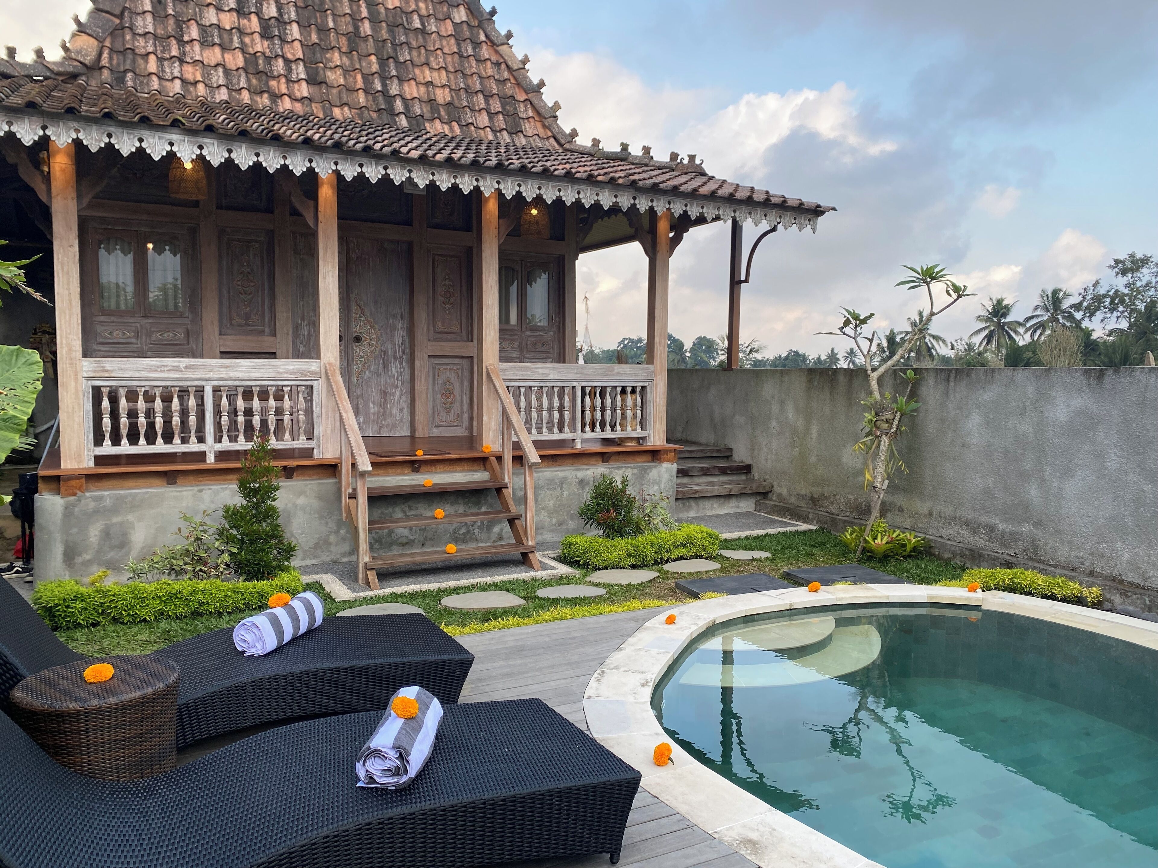 A traditional wooden house with a tiled roof, a covered porch, and a pool area with lounge chairs and a small garden.