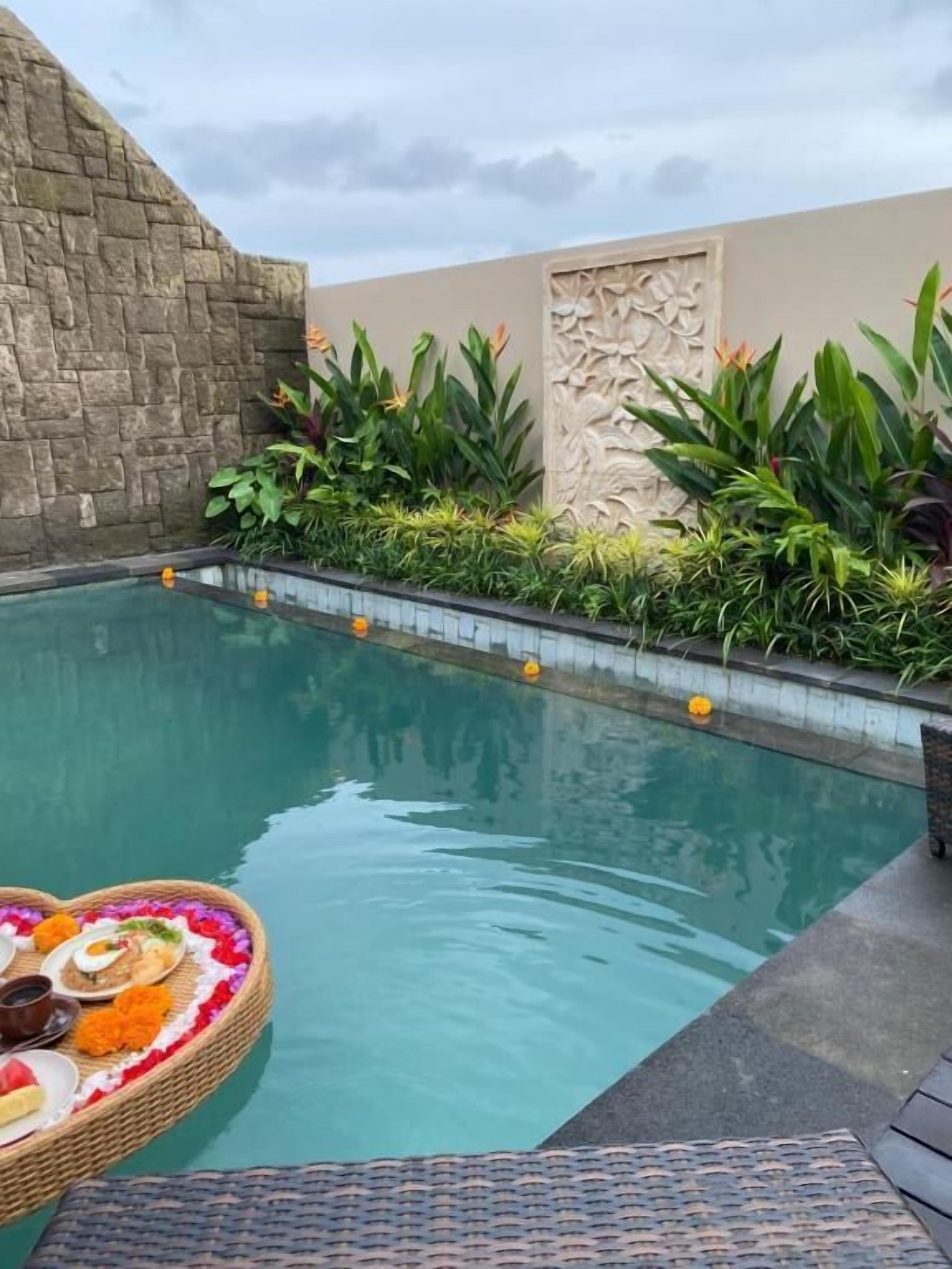 A swimming pool with a heart-shaped floating basket containing food and flowers, surrounded by lush greenery and a stone wall.