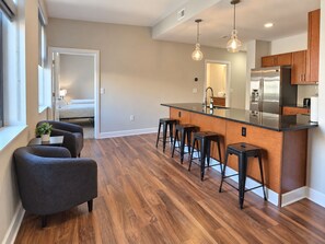 A modern kitchen with a central island, bar stools, wooden cabinets, and a view into the adjacent bedroom. - Downtown Suite - Spacious! RnRHoF, Casino, Stadium (Cleveland)