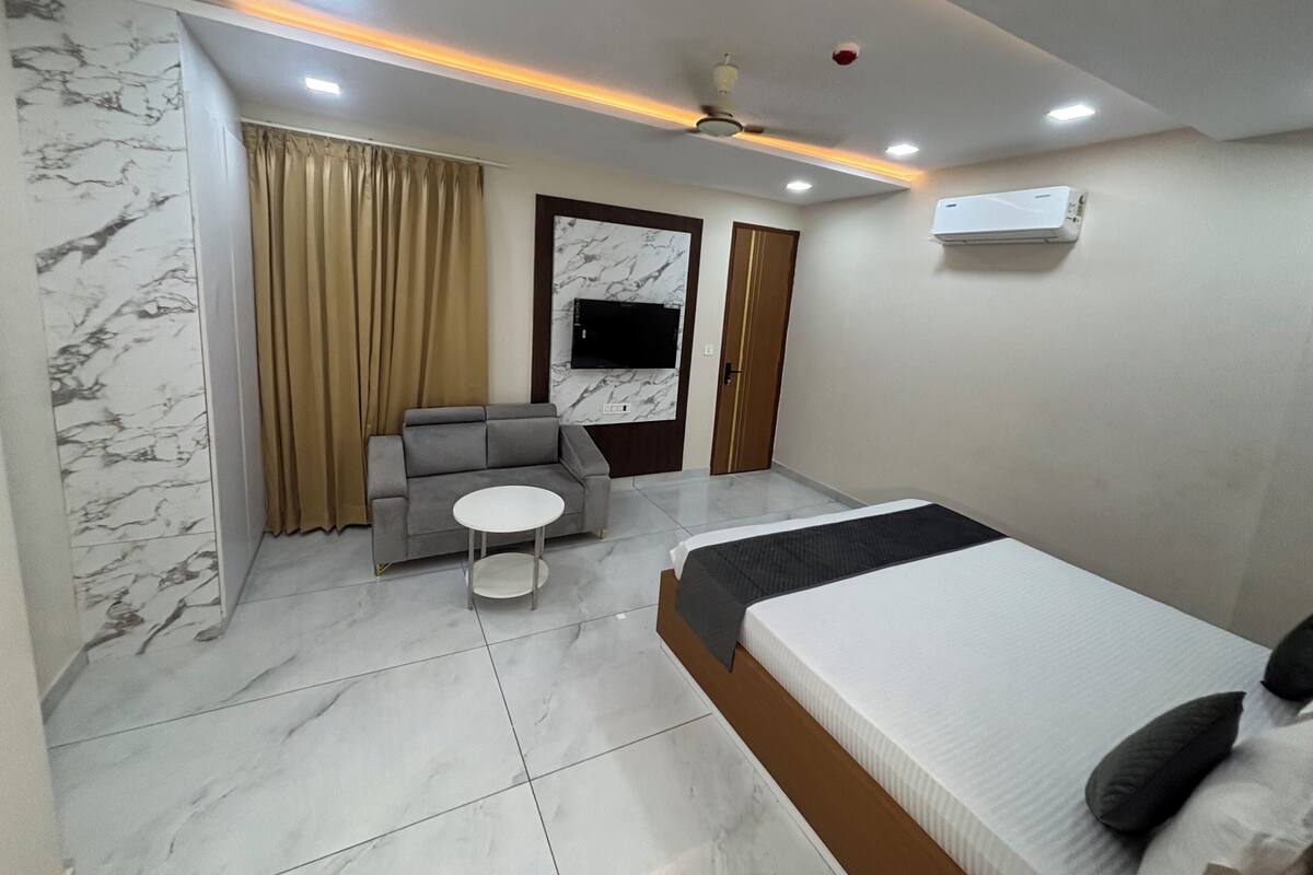 A modern hotel room with a bed, a sofa, a small round table, and a TV mounted on the wall.