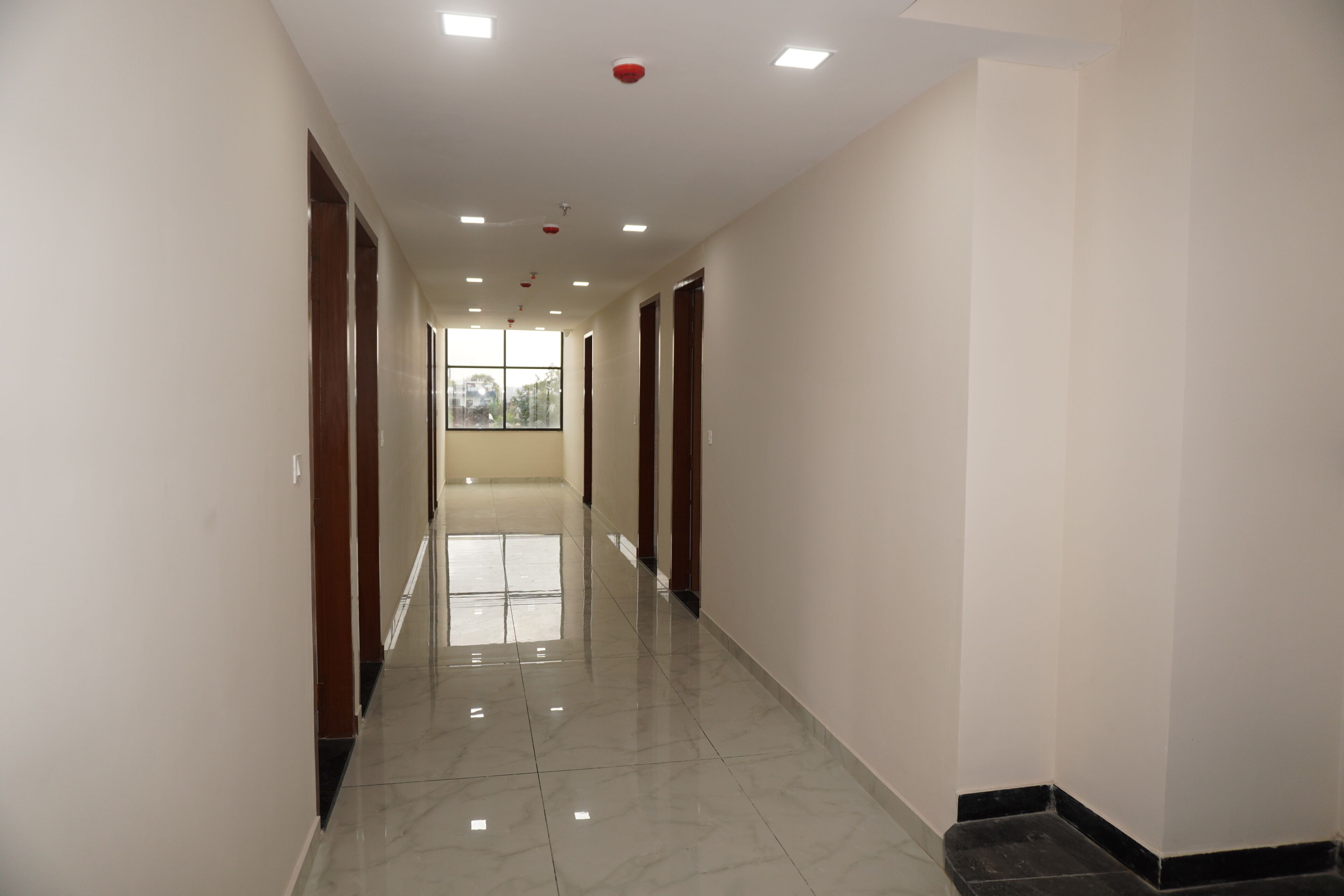 A clean, well-lit hallway with a shiny tiled floor, wooden doors, and a window at the end.