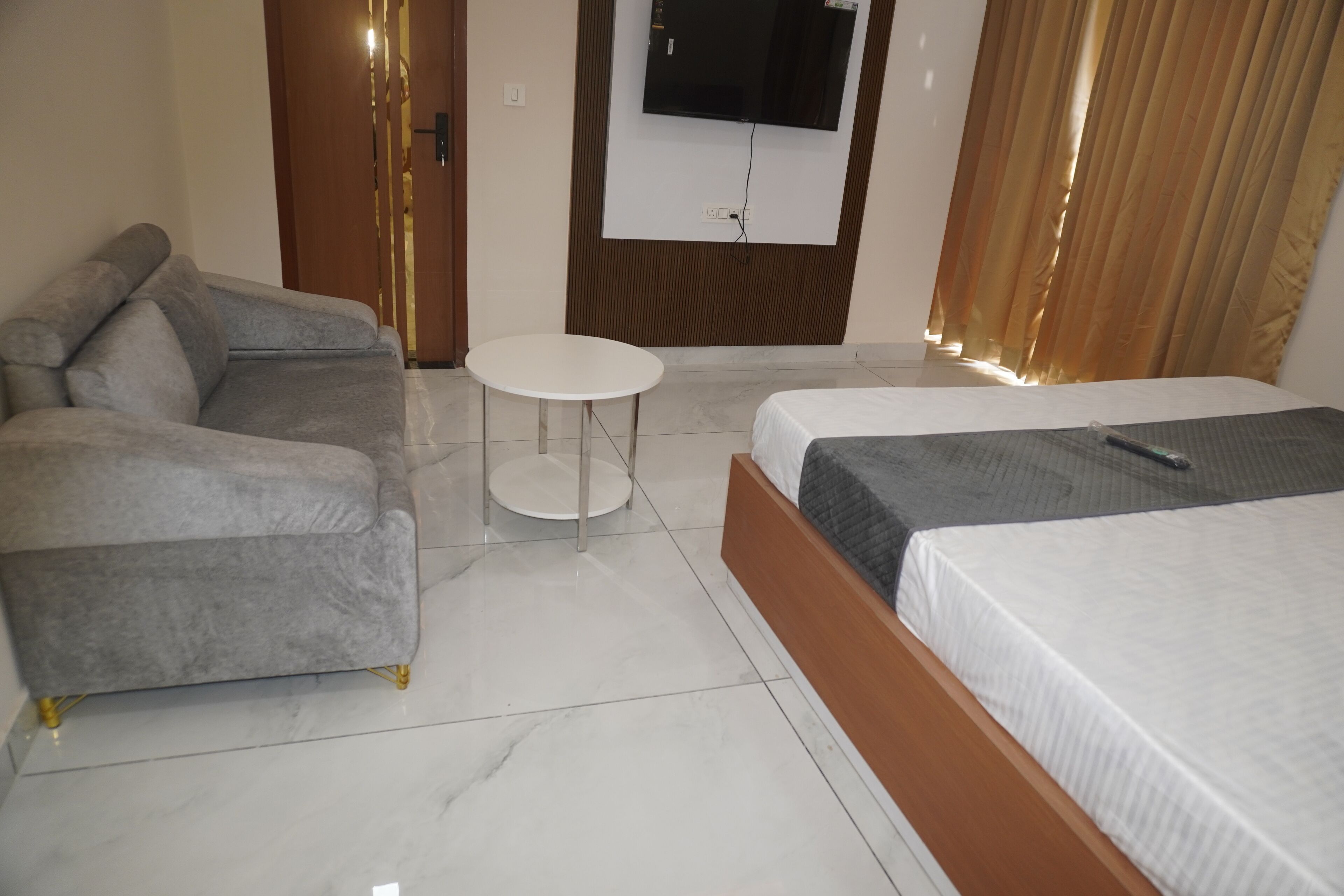 A modern hotel room with a grey sofa, a round white table, a bed with a grey blanket, and a TV mounted on the wall.