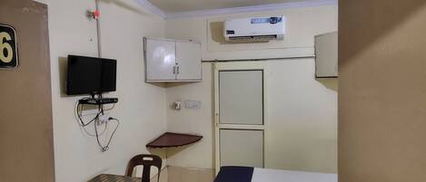 Una habitación de hotel con cama, escritorio, silla, televisor, equipo de aire acondicionado y ventilador de techo.