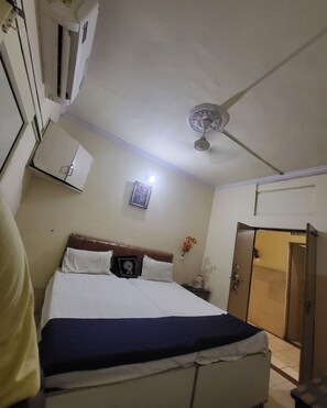 Hotellihuone, jossa on sänky, kattotuuletin, ilmastointilaite, ovi ja pieni pöytä, jossa on lamppu.
