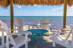 Een houten terras met witte Adirondack-stoelen en een turquoise tafel, met uitzicht op een rustige zee.