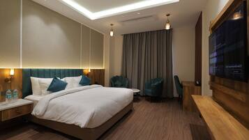 A modern hotel room with a large bed, two armchairs, a wooden desk, and a flat-screen TV.