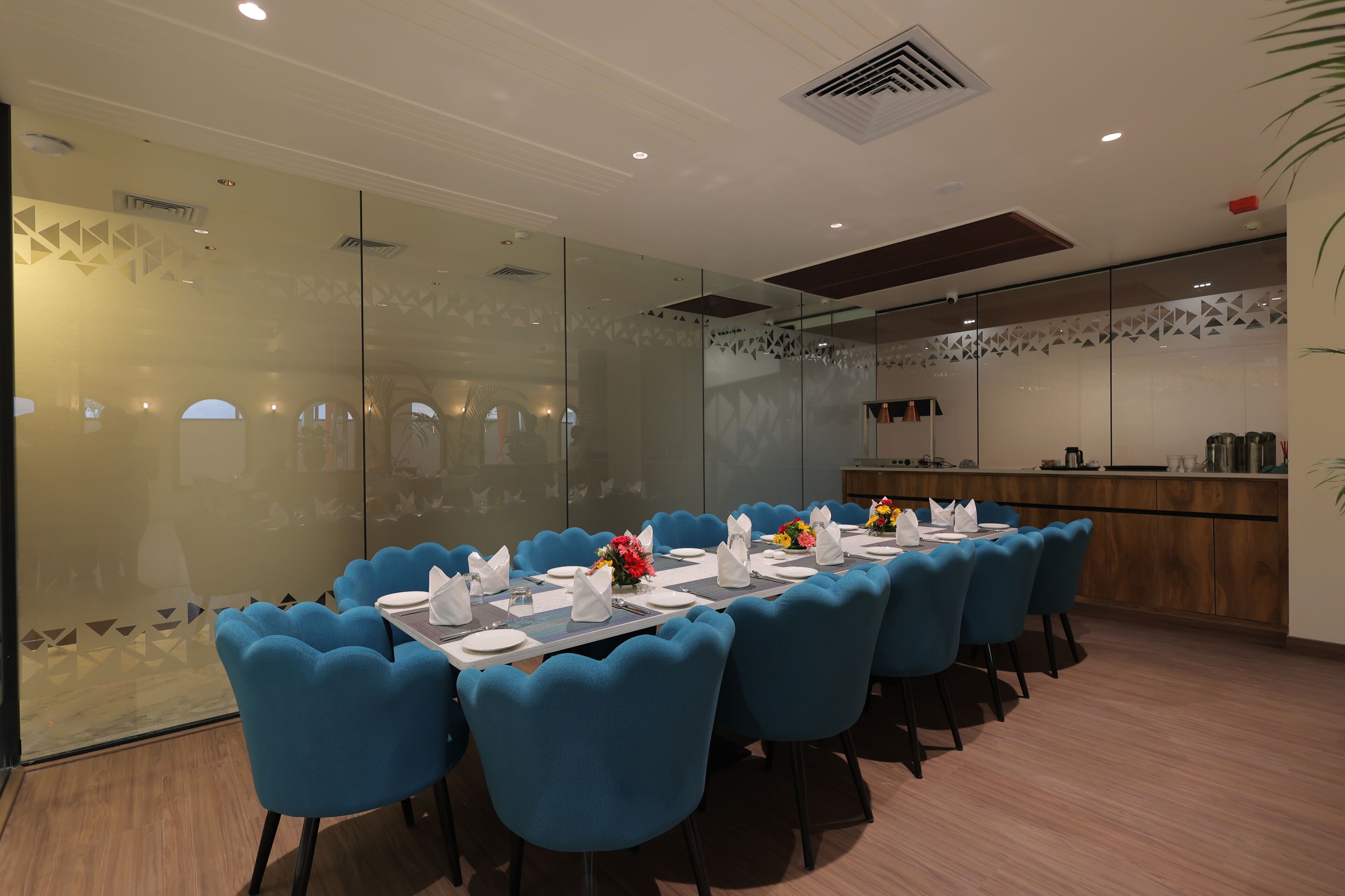 A conference room with a long table set for a meeting, blue chairs, and a kitchen area in the background.