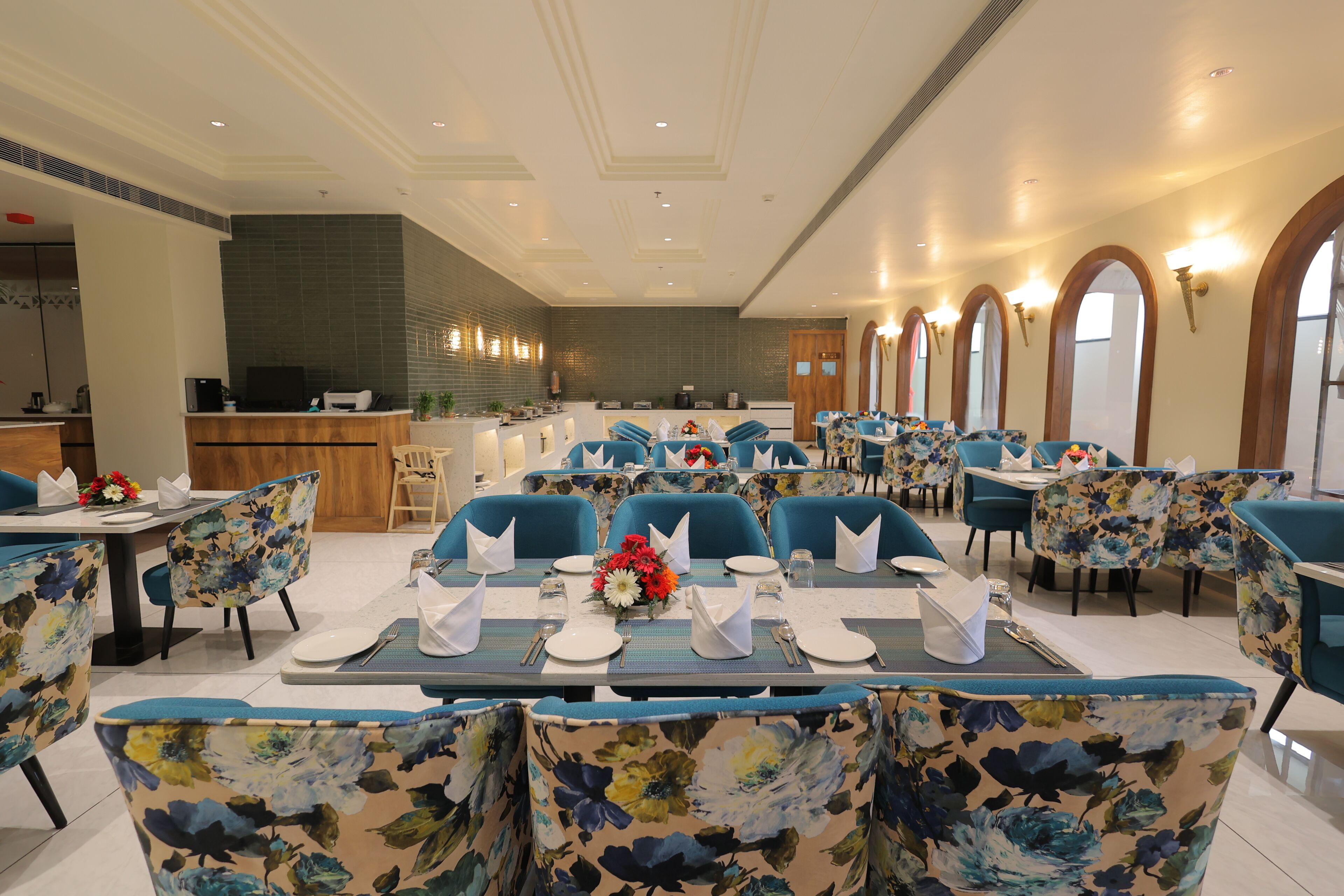A dining area with tables set for service, blue floral chairs, and a kitchen in the background.
